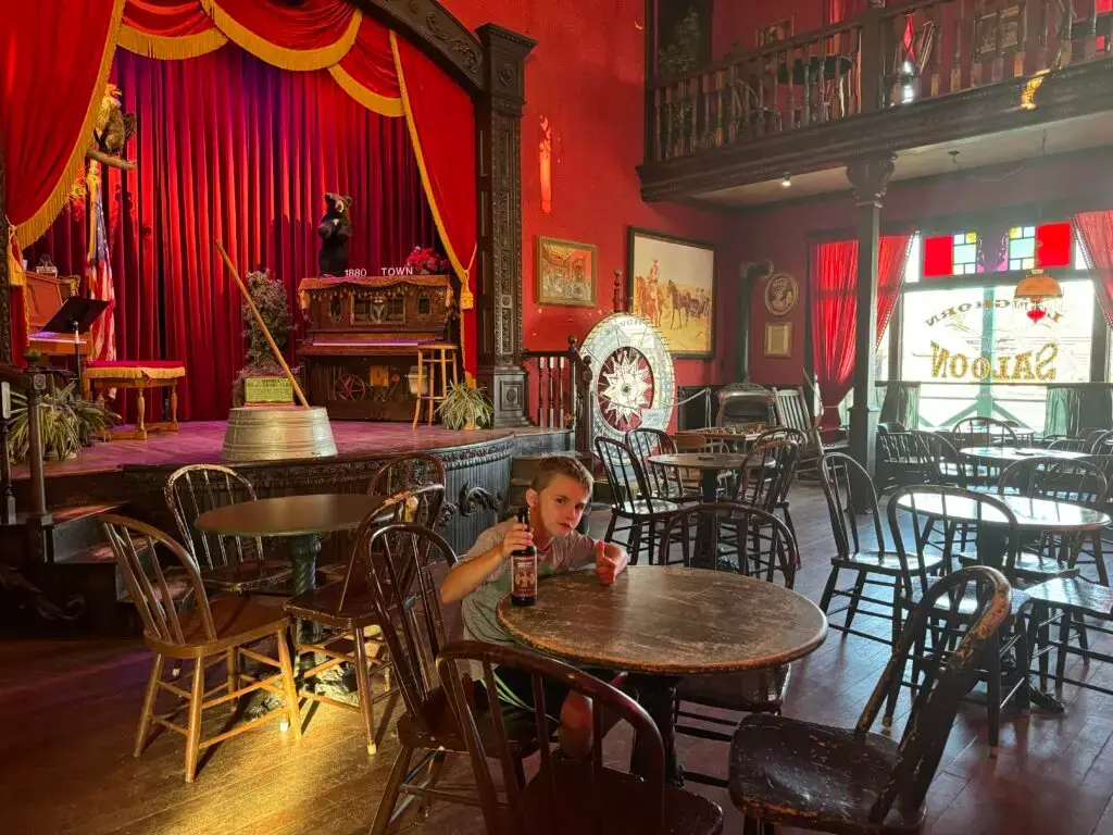 A child sits at a table with a drink in an old-fashioned saloon, featuring a stage with red curtains and vintage decor. 1880 Town with kids