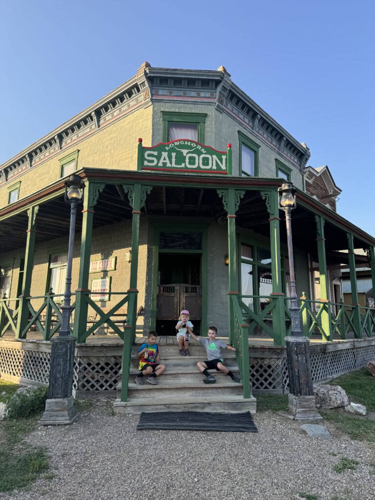 Three children sit on the steps of a green saloon with a wraparound porch under a clear blue sky. 1880 Town with kids