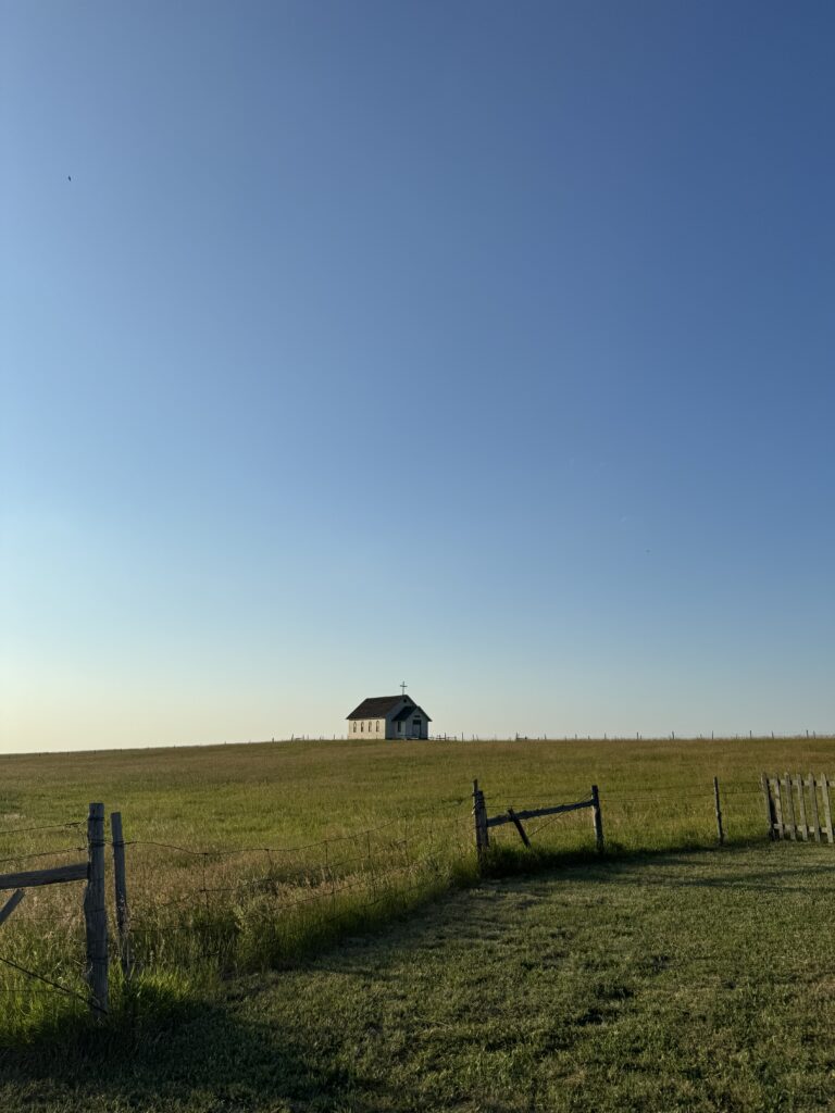 A solitary building stands in the middle of a vast grassy field under a clear blue sky. A wooden fence is in the foreground. 1880 Town with kids