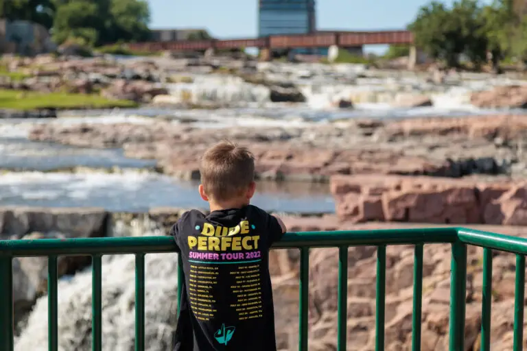 A child wearing a "Dude Perfect Summer Tour 2022" shirt stands at a railing, overlooking a river with rocky sections and a bridge in the background. Sioux Falls with Kids