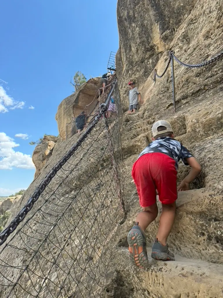 A child climbs stone steps, secured by chains and netting, against a rocky cliff under a blue sky at Mesa Verde. Other kids are visible ahead, making for an adventurous family outing amidst the ancient wonders.