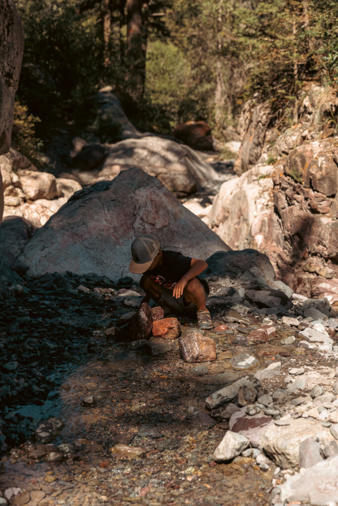 A child crouches beside a shallow stream in a rocky, wooded area, examining the rocks. 2 days in Ouray with kids.