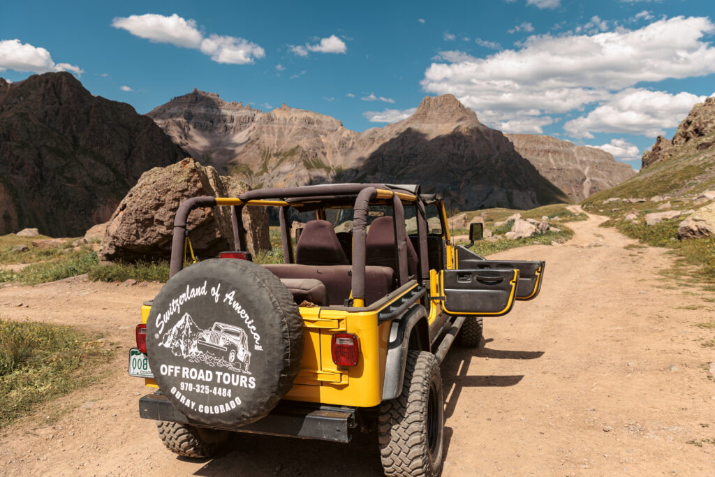 Yellow off-road vehicle with an open roof on a rocky dirt trail surrounded by mountains under a blue sky. Spare tire on the back reads "Off Road Tours.  2 days in Ouray with kids.