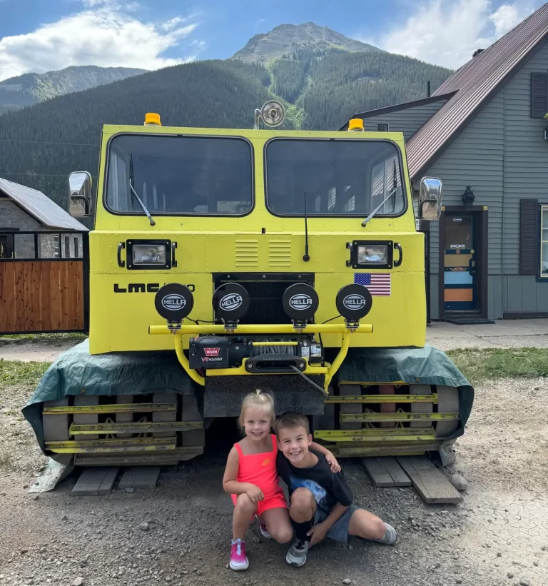 Two children sit under a large yellow snow vehicle parked on wooden planks, with mountains and houses in the background. Afternoon in Silverton with Kids.