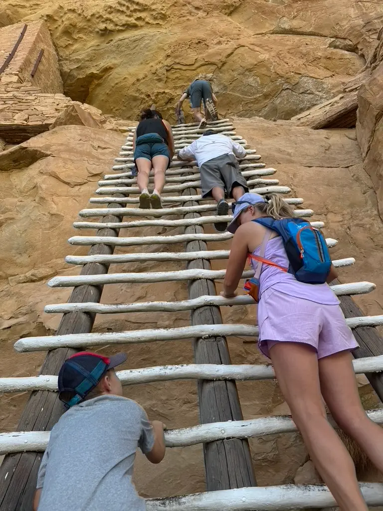 People climbing a steep wooden ladder on a rocky cliff, with a person wearing a backpack in the foreground, captures the adventurous spirit of exploring Mesa Verde with kids.