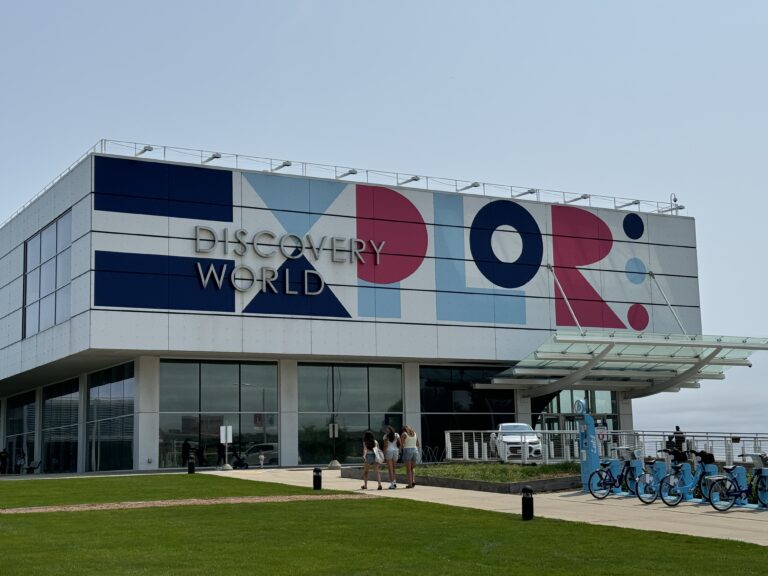 Modern building labeled "Discovery World" with "EXPLORE" on the facade. People walk nearby, and bicycles are parked under a glass canopy.