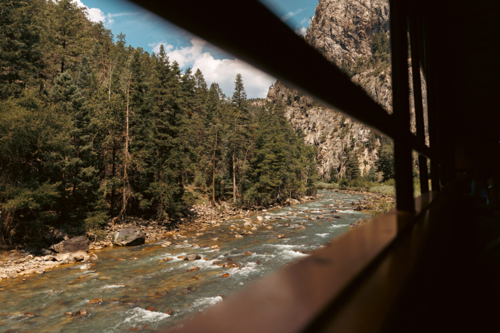 From the window of the Durango train with kids, watch as a flowing river winds its way past dense green trees and rugged rocky cliffs, all under a partly cloudy sky.