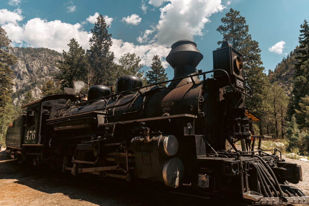 A vintage steam locomotive, reminiscent of the Durango train, sits on the tracks surrounded by trees and hills under a partly cloudy sky, perfect for an adventure with kids.  Durango train with kids.