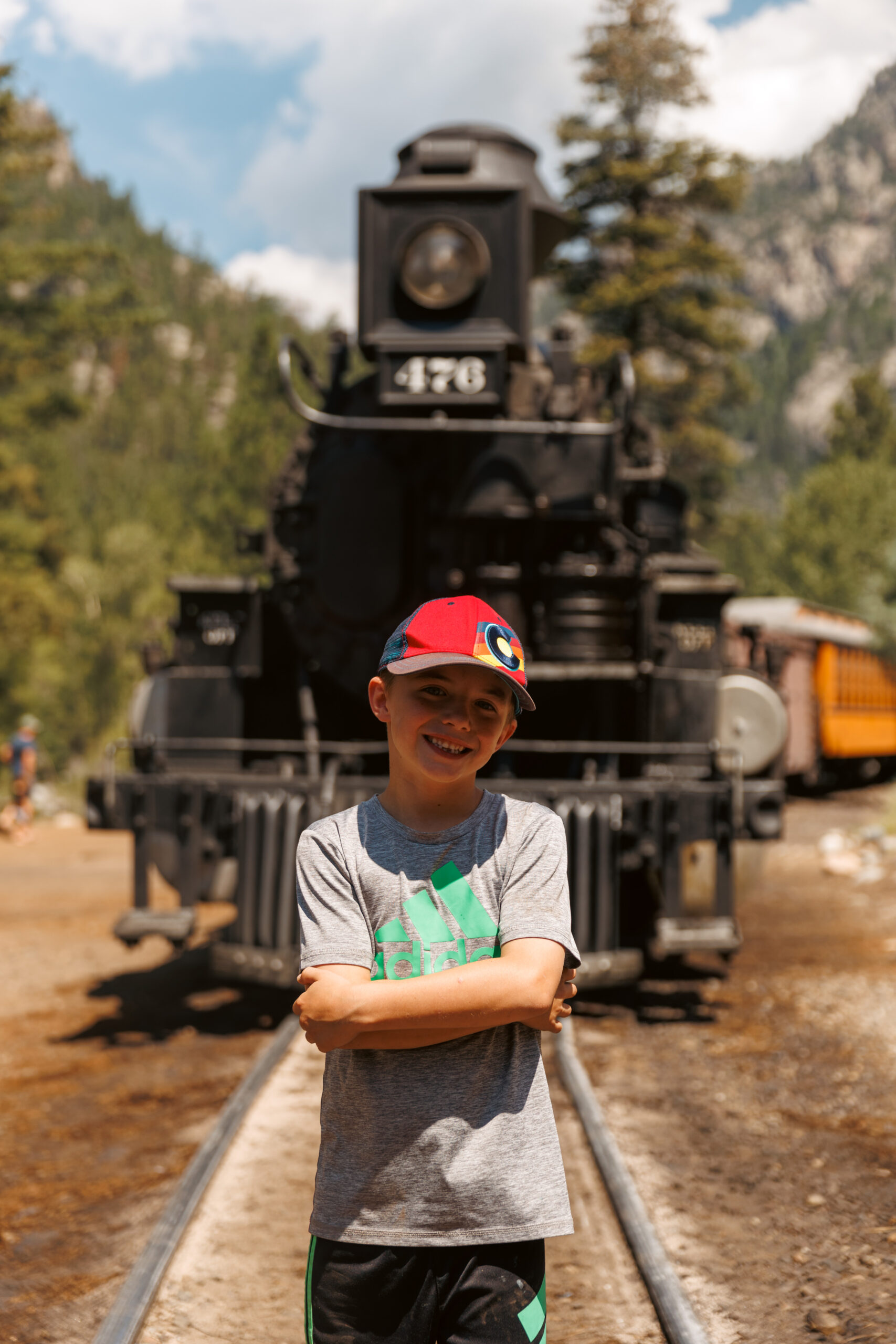 A young boy in a gray shirt and red cap stands on train tracks with an old steam locomotive and trees in the background.  Durango train with kids.