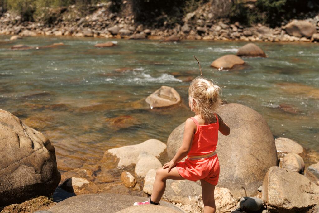 Child in a pink outfit stands on a large rock by a flowing river, holding a stick, with rocky riverbank in the background.  Durango train with kids.