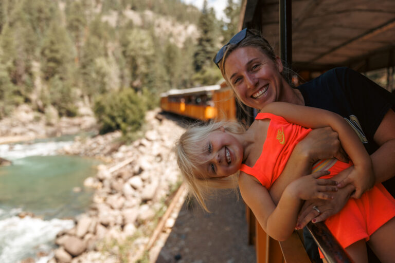 A woman beams while holding a smiling young girl in an orange outfit aboard the Durango train, enjoying the scenic view of a river and forest. Durango train with kids.