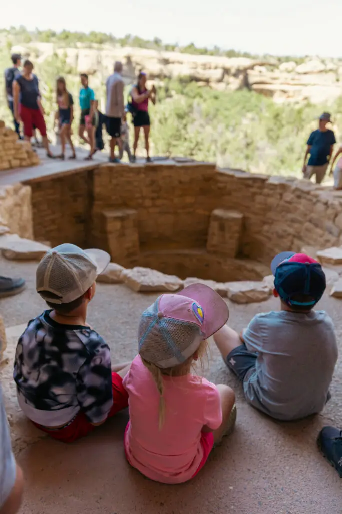 Three children sit on the ground, facing a circular stone ruin at Mesa Verde. Other people stand in the background at a higher level, exploring the historical site's rich tapestry with kids. The setting appears calm and inviting amidst ancient surroundings.