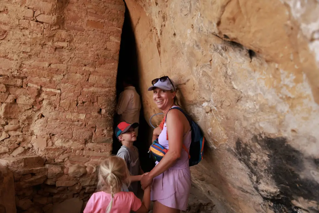 A woman holds a child's hand while standing in a narrow rocky passageway, with another child nearby. They appear to be exploring the fascinating ancient ruins of Mesa Verde with kids, embarking on an adventure through the echoes of history and timeless landscapes.