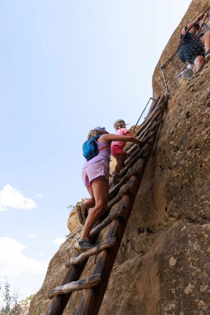 Two people climb a wooden ladder against a rocky surface under a clear sky, exploring the stunning landscape of Mesa Verde with kids.