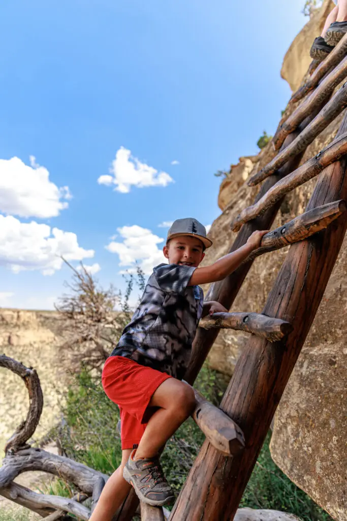 A child scales a wooden ladder against a rugged rock face under a blue sky dotted with clouds, embodying the adventurous spirit of exploring Mesa Verde with kids.