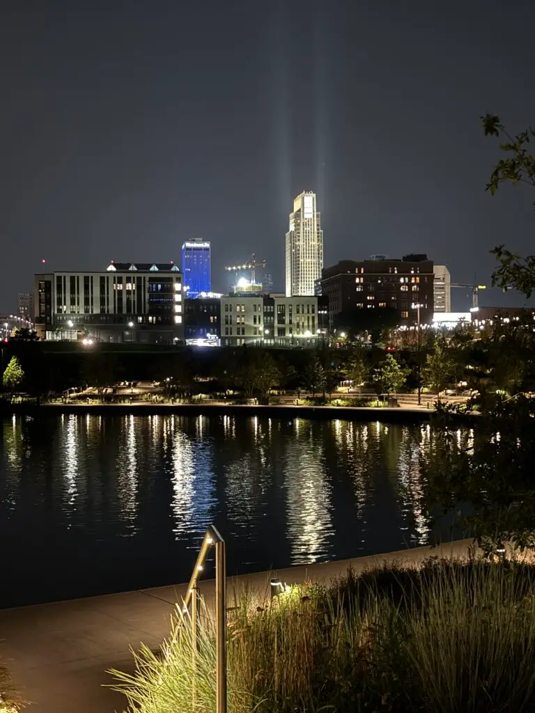 Nighttime cityscape with buildings lit up, reflecting on a calm river in the foreground, surrounded by greenery. Omaha with kids