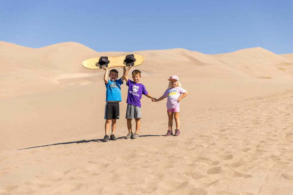 Three children stand on the Great Sand Dunes; the boy in the middle hoists a sandboard overhead. Clad in casual summer clothes, they pose under a clear blue sky, ready for an adventure.